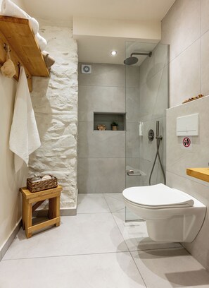 Une salle de bain moderne avec un mur d’accent en pierre, une douche en verre, un tabouret en bois avec un panier et des toilettes blanches.