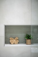Un baño con pared de azulejos blancos, cerramiento de ducha de vidrio y una repisa con una cesta tejida y una planta en maceta.
