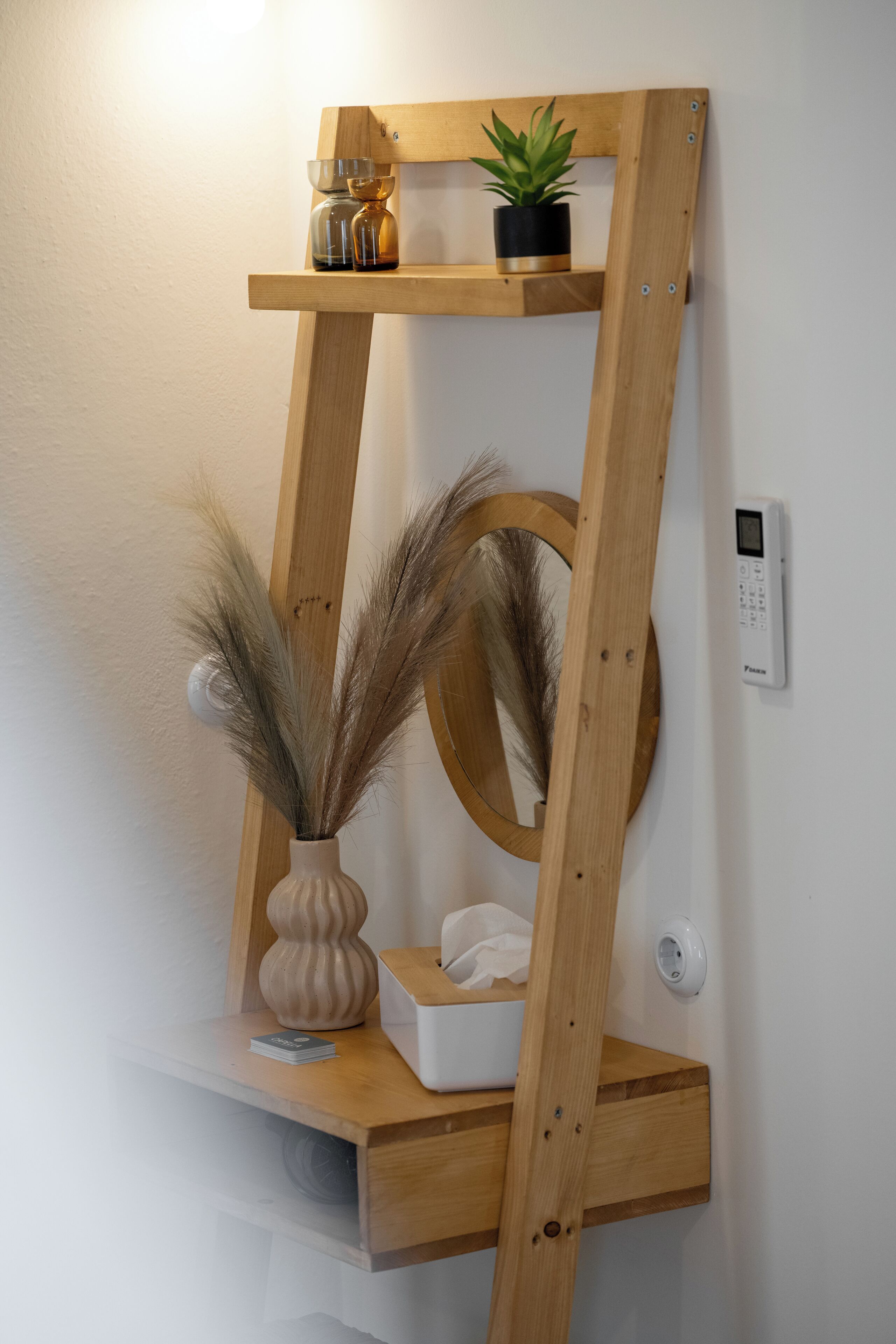En træreol med et spejl, en vase med pampas, en lille potteplante og en æske med vatrondeller.