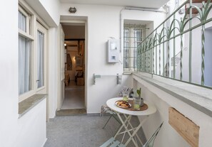 Un couloir étroit avec une petite table et des chaises, une plante en pot et une fenêtre ornée d’une rambarde en métal.