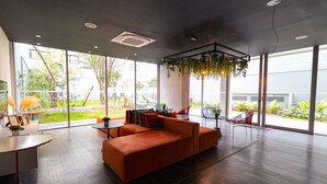 A spacious room with a large orange sectional sofa, a small round table, and a chair. There are large windows providing a view of the outdoors. - Vossa Bossa Moema ON (São Paulo)