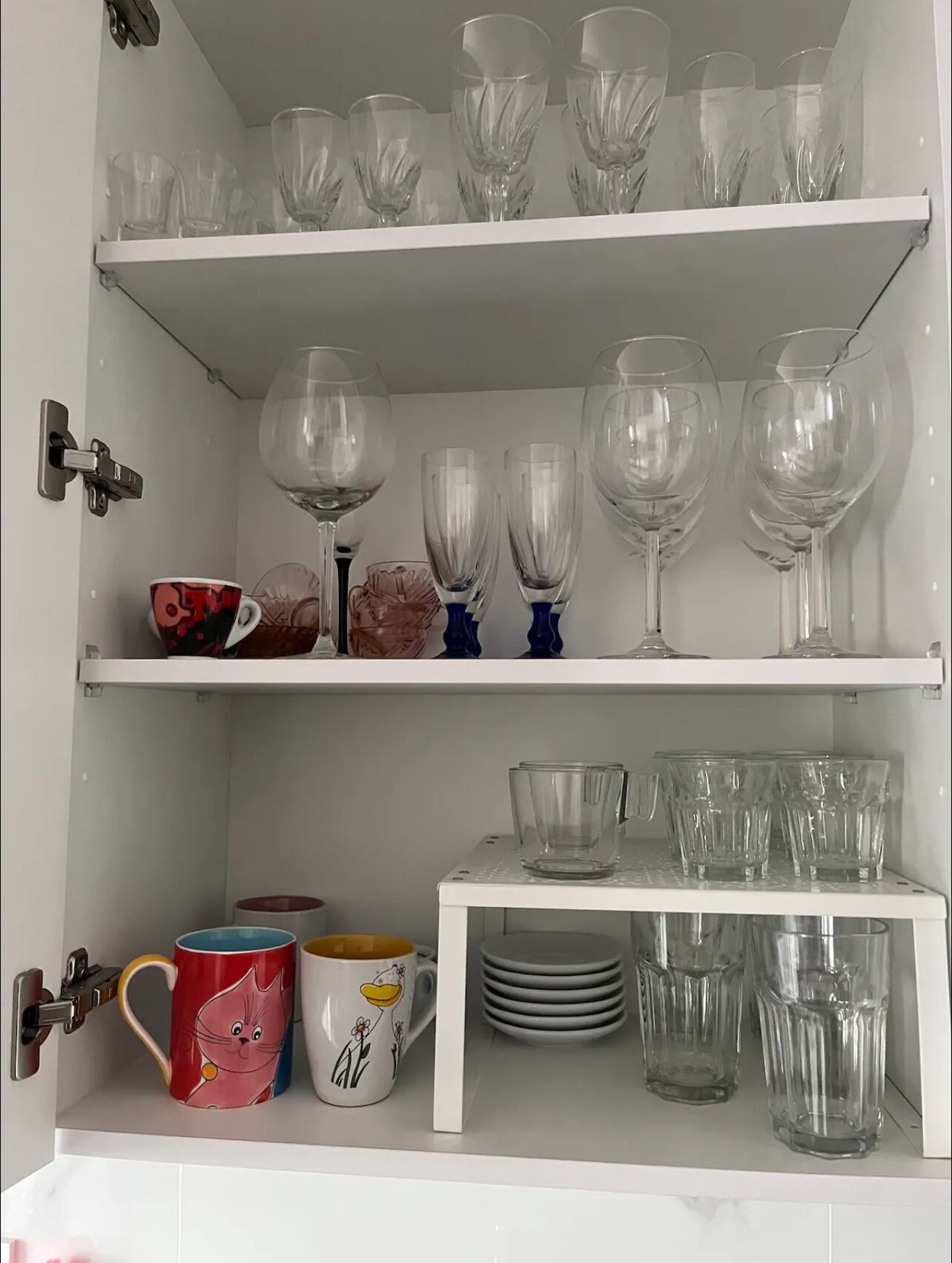Ein weißes Schränkchen mit Regalen, auf denen verschiedene Gläser, darunter Weingläser und Tumbler, stehen.