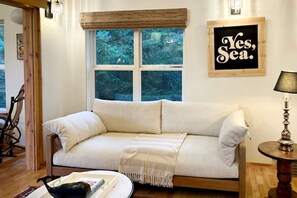 A cozy room with a white sofa, a wooden rocking chair, a small table with a lamp, and a window with a view of trees.