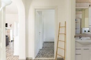 Un pasillo con una puerta blanca, una escalera de madera y un baño con lavamanos y espejo.