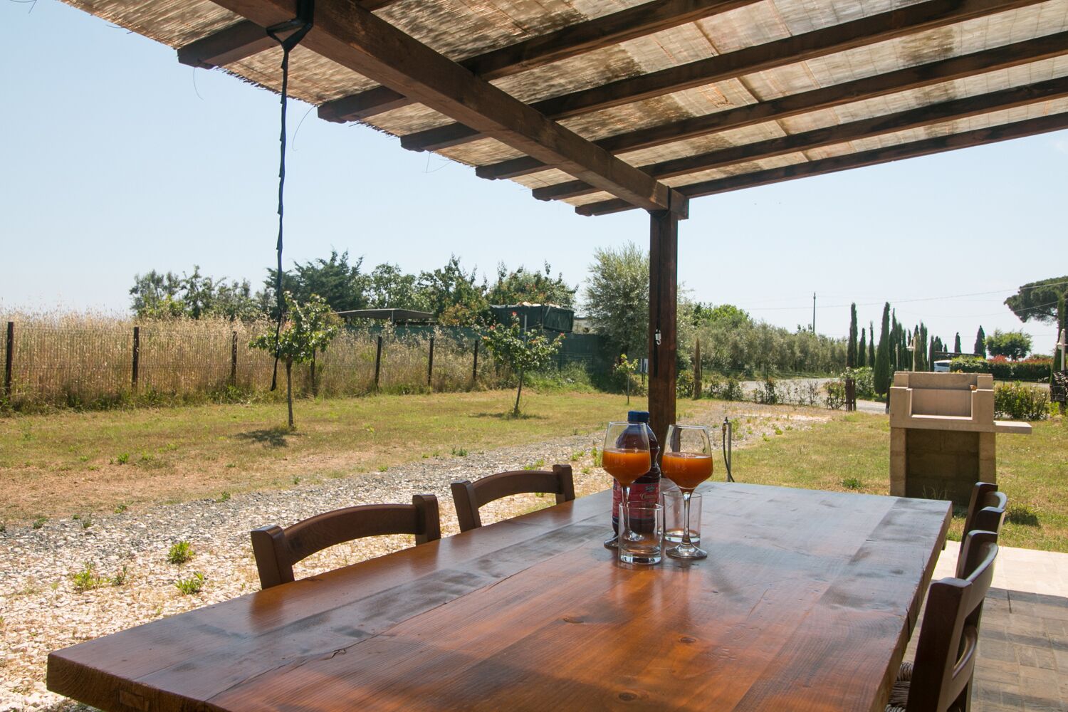 An outdoor wooden table with chairs, a bottle of orange juice, and a glass, set under a pergola with a clear view of a grassy area and trees in the background.