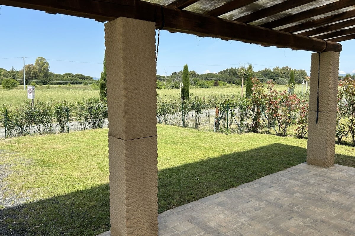 Un espacio exterior techado con pilares de piedra, suelo empedrado y vistas a un campo cubierto de césped, plantas y árboles.