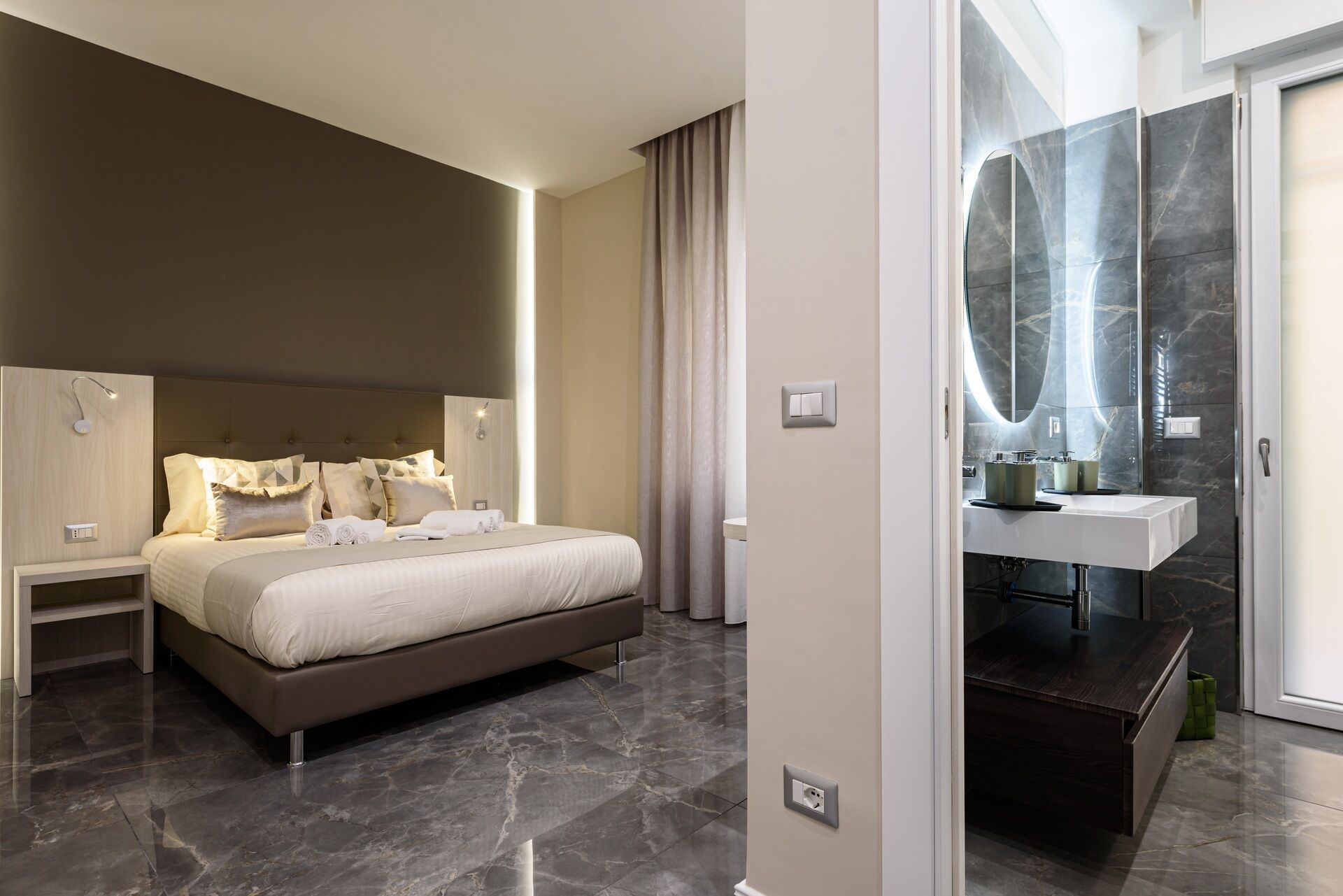 Une chambre d’hôtel moderne avec un grand lit, des tables de chevet et une salle de bain dotée d’un lavabo et d’un miroir en marbre.