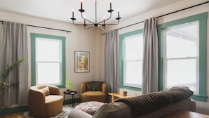 Un salon chaleureux avec un canapé, un fauteuil et une petite table sur laquelle est posée une plante. De grandes fenêtres, ornées de rideaux, laissent entrer la lumière, et un lustre illumine la pièce.