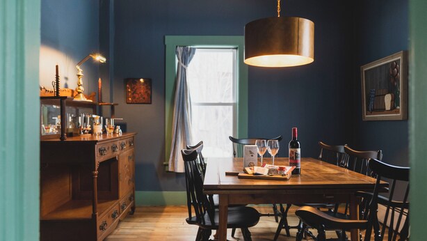 Un coin repas comprenant une table en bois, des chaises, un lustre, une fenêtre avec des rideaux et un meuble contenant des bouteilles.