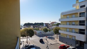 A street view with a building featuring balconies and a clear view of the sea in the background. - Praia da Saude (Setúbal)
