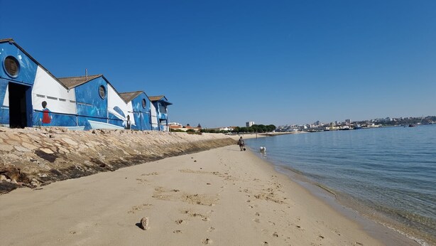 A beach with a stone wall and blue buildings with white accents. - Praia da Saude (Setúbal)