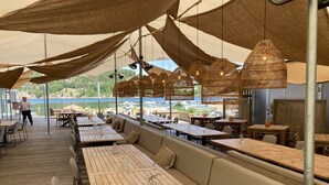 Un restaurante al aire libre con mesas y bancos de madera, vista al agua y un toldo.