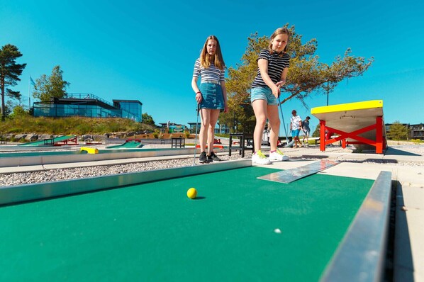 Two people playing a game on a green table with a yellow ball, a red and yellow structure, and a clear blue sky in the background.