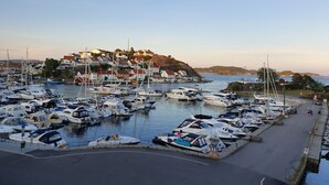 Un puerto deportivo con numerosos barcos amarrados, un pequeño pueblo con casas de tejados rojos en una colina, y un cielo despejado.