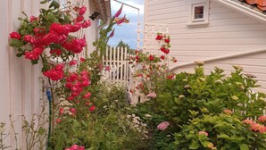 Una casa blanca con techo de tejas rojas, cerca de piquetes blancos y un jardín con flores rosas.