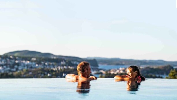 Dos personas en una piscina infinita con vistas a un pueblo en la ladera.