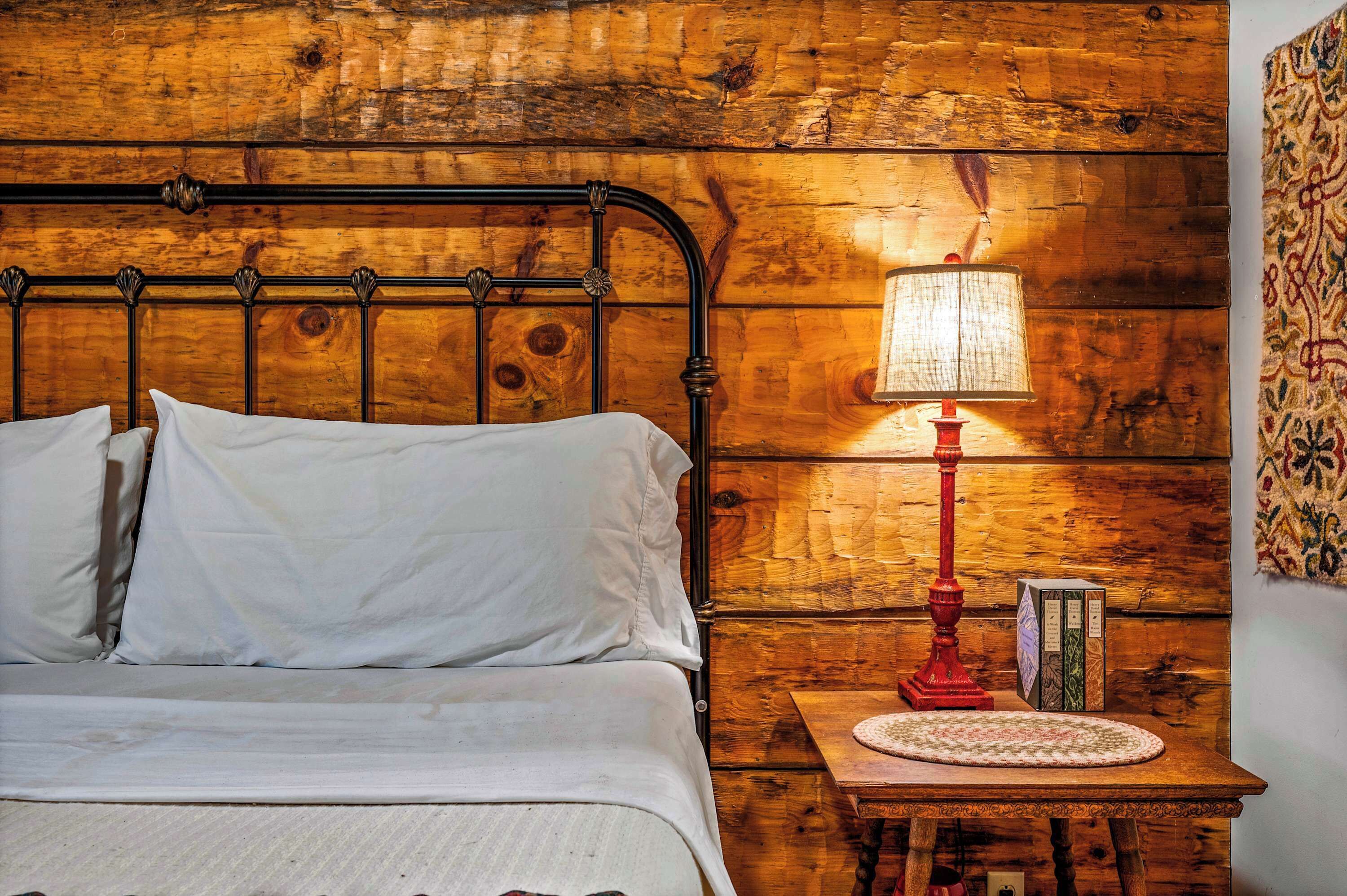 Ein Bett mit weißen Leinen, einem Metall-Kopfschild, einer roten Tischlampe und einem kleinen Nachttisch mit Büchern.