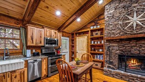 A wooden cabin interior with a stone fireplace, wooden cabinets, a dining table, and a kitchen area.