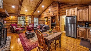 A log cabin interior with a dining area, wooden furniture, and a kitchen with a stainless steel refrigerator.