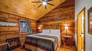 A wooden cabin bedroom with a bed, two bedside lamps, a ceiling fan, and a window with curtains.
