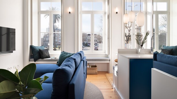 Una sala de estar moderna con un sofá azul, un televisor de pantalla plana y ventanas amplias que ofrecen vistas a la ciudad.