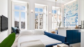 Un dormitorio moderno con una cama grande, un sofá azul, una mesita pequeña y vistas de la ciudad a través de amplias ventanas.