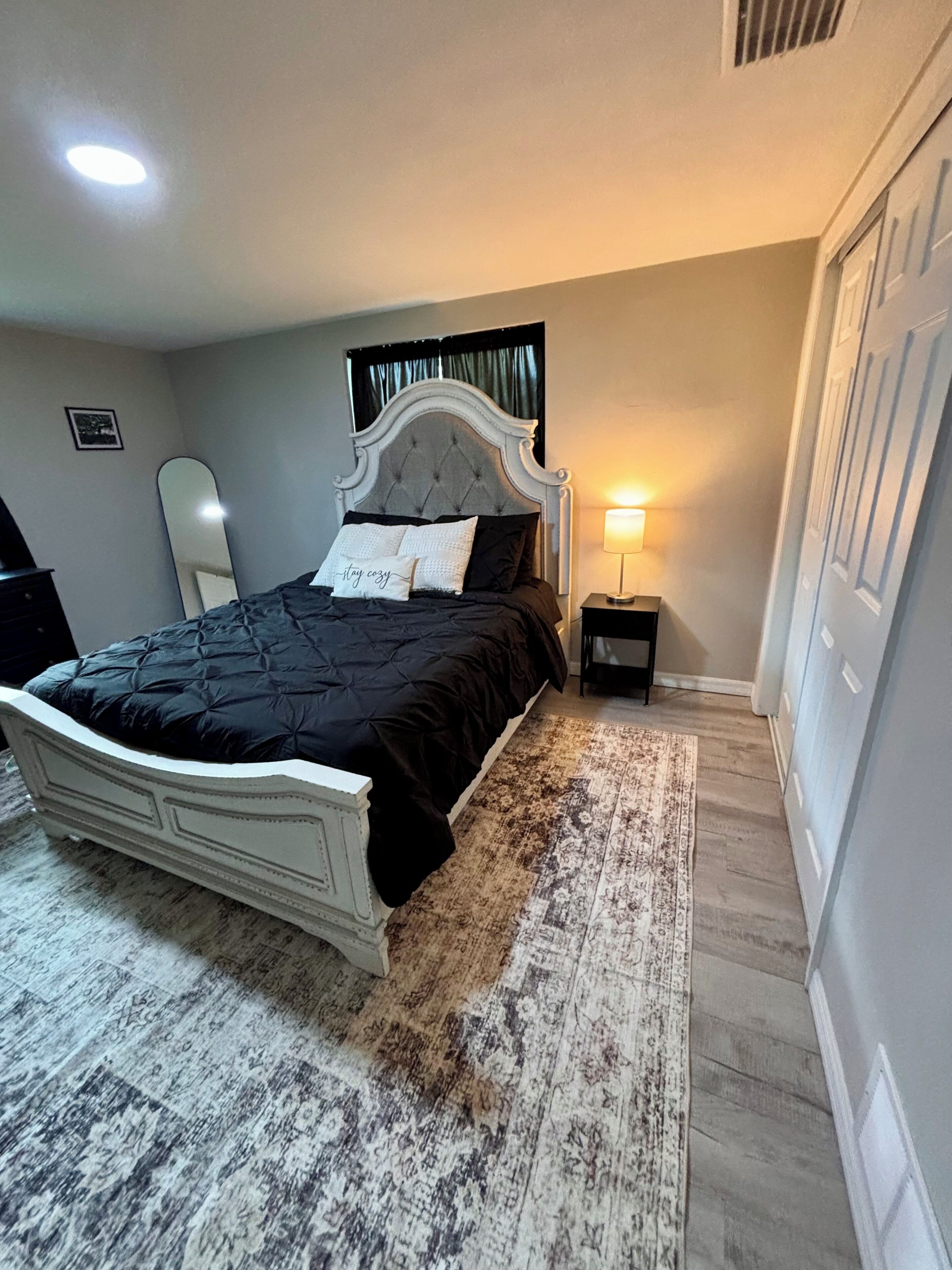 A bedroom with a bed, a bedside table, a lamp, a mirror, and a door.
