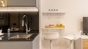 A modern kitchen with a stainless steel sink, a double faucet, and a wooden tray with croissants and orange juice. There are two white bar stools and a white shelf above the counter. - Charming Studio for Two in Beautiful Chamberí (Madrid)