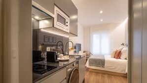 A compact kitchenette with a microwave, stove, and washing machine, a small dining area with a table and chairs, and a bedroom with a bed and pillows. - Charming Studio for Two in Beautiful Chamberí (Madrid)