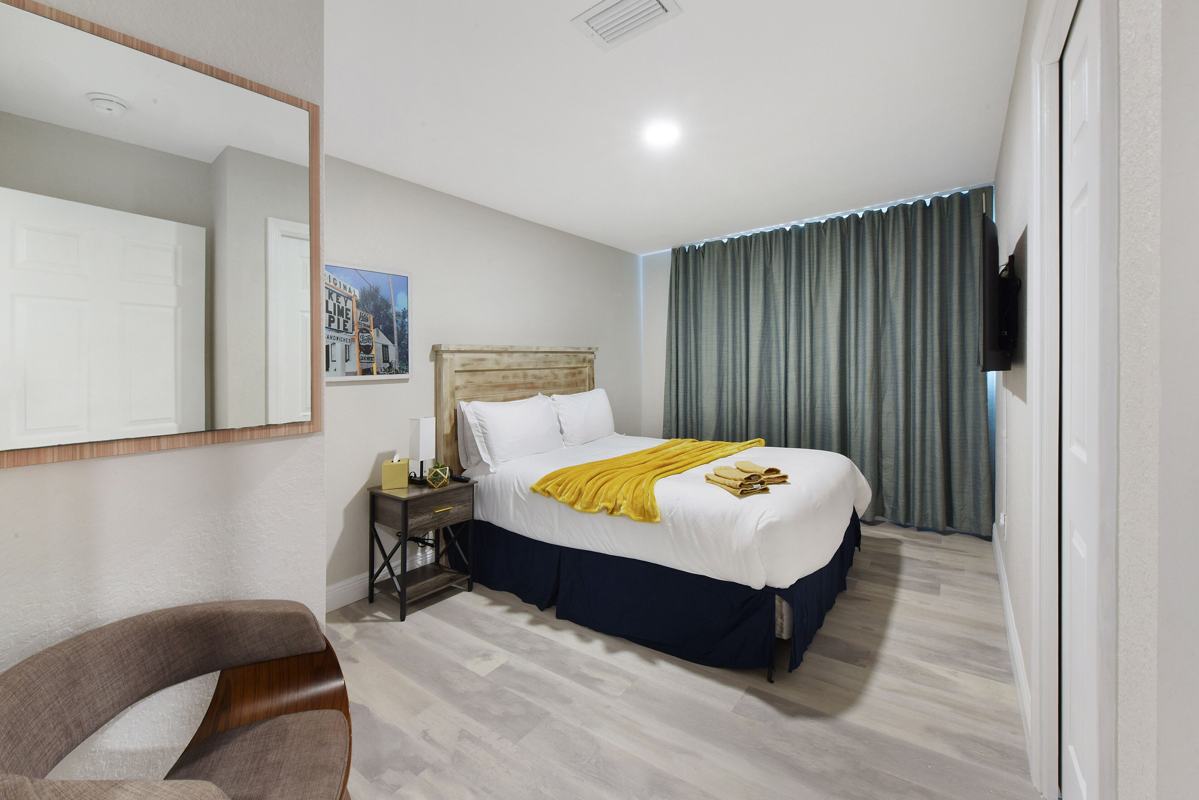Ein Hotelzimmer mit Bett, Nachttisch, Stuhl, Spiegel und einem Fenster mit Vorhängen.