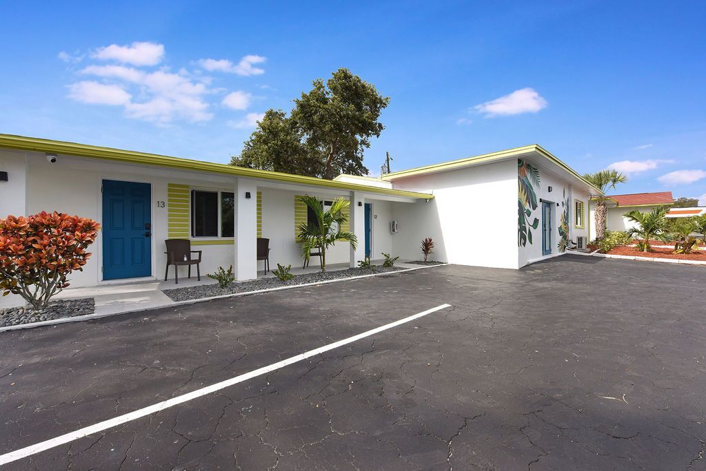 Un motel con aparcamiento pavimentado, una puerta azul y un pequeño jardín con plantas y una silla.