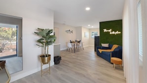 Un salon moderne avec un coin repas, une grande plante en pot, un canapé bleu et un panneau lumineux au néon sur le mur.