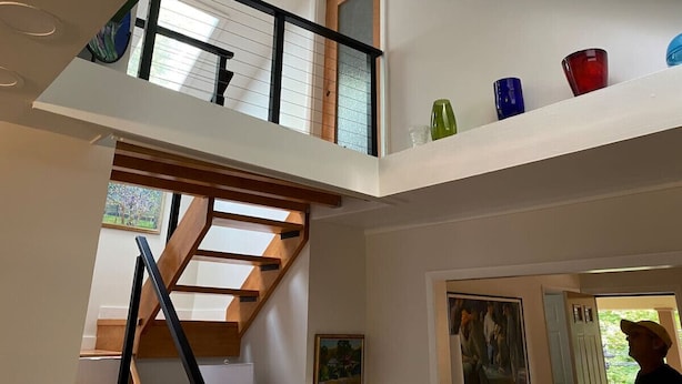 Un interior de varios niveles con una escalera de madera, un balcón con objetos de vidrio de colores y un cuadro en la pared.