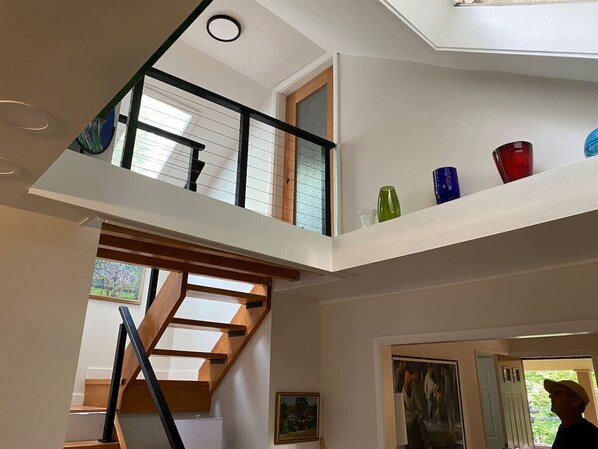 Un intérieur à plusieurs niveaux avec un escalier en bois, un balcon agrémenté d’objets en verre aux couleurs vives et un tableau accroché au mur.