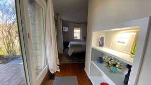 Un couloir étroit avec un plancher en bois, un lit à l’arrière-plan et une étagère ornée d’objets décoratifs.