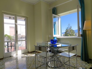A room with a glass table, chairs, a blue bowl, a window with curtains, and a balcony with a railing.