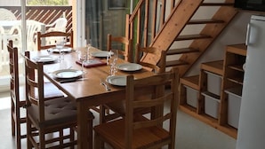 Un comedor con una mesa de madera dispuesta para cuatro personas, rodeada de sillas de madera. Se ve una escalera con barandillas de madera al fondo.