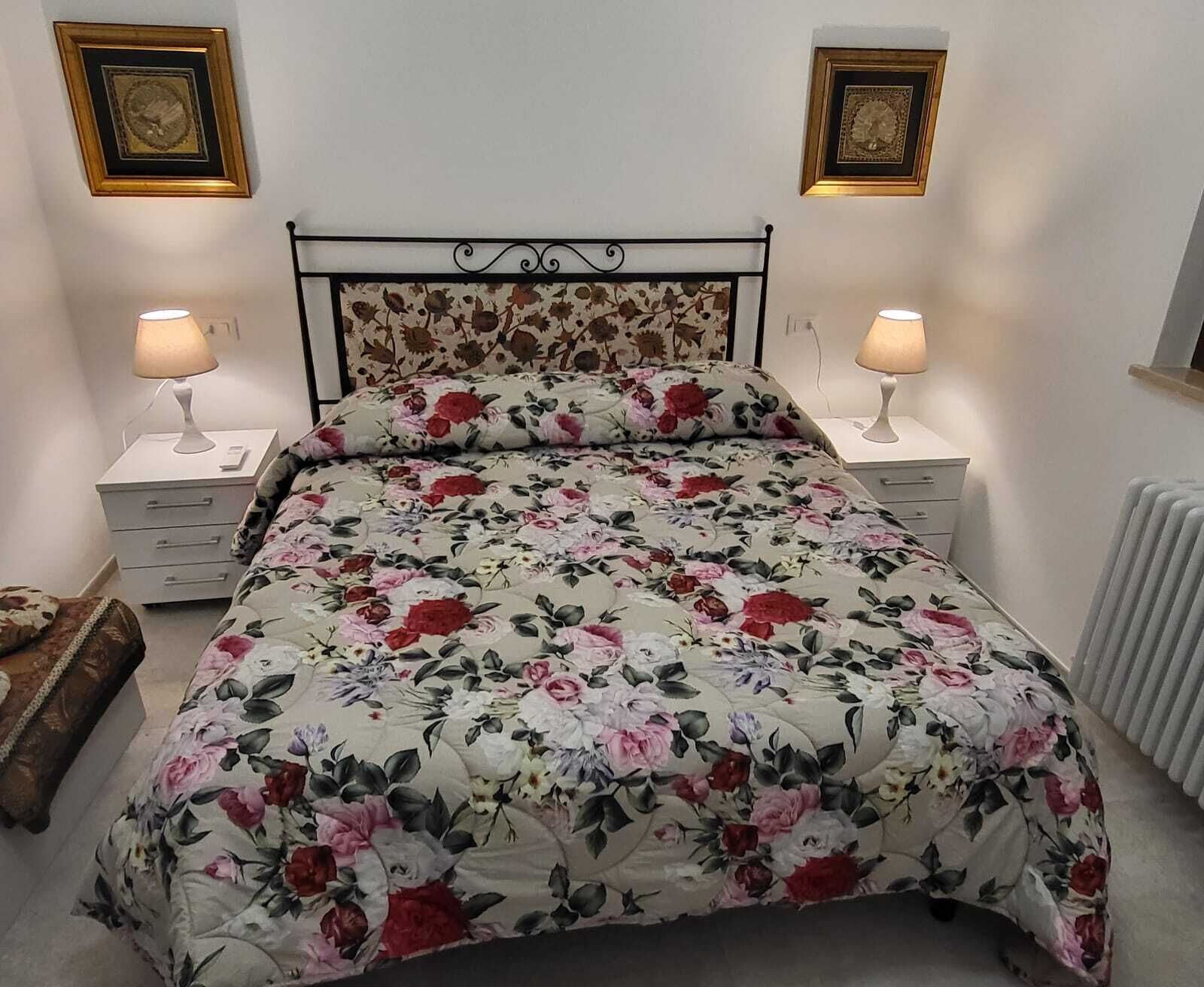 Una cama con colcha estampada, dos mesitas de noche con lámparas, un radiador y dos cuadros en la pared.