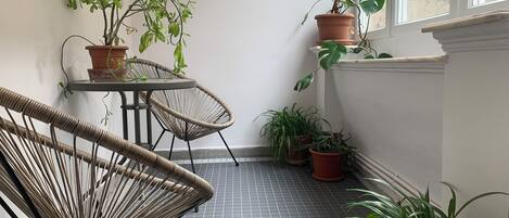 A corner room with a tiled floor, a wicker chair, a small table, and potted plants.