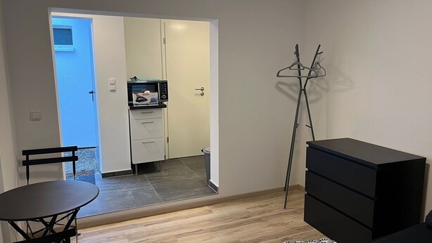 Une pièce compacte comprenant une table à manger, une commode noire et un porte-manteau. Une porte donne sur une petite cuisine équipée d’un four à micro-ondes et d’une petite fenêtre.