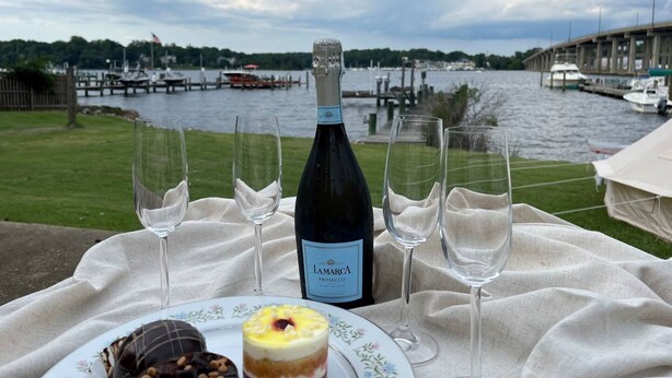 A table set with a bottle of wine, glasses, and a plate with food, overlooking a body of water with boats and docks. - Dock, Dine, & Unwind: The Ultimate Waterfront Glamp Spot (Edgewater)