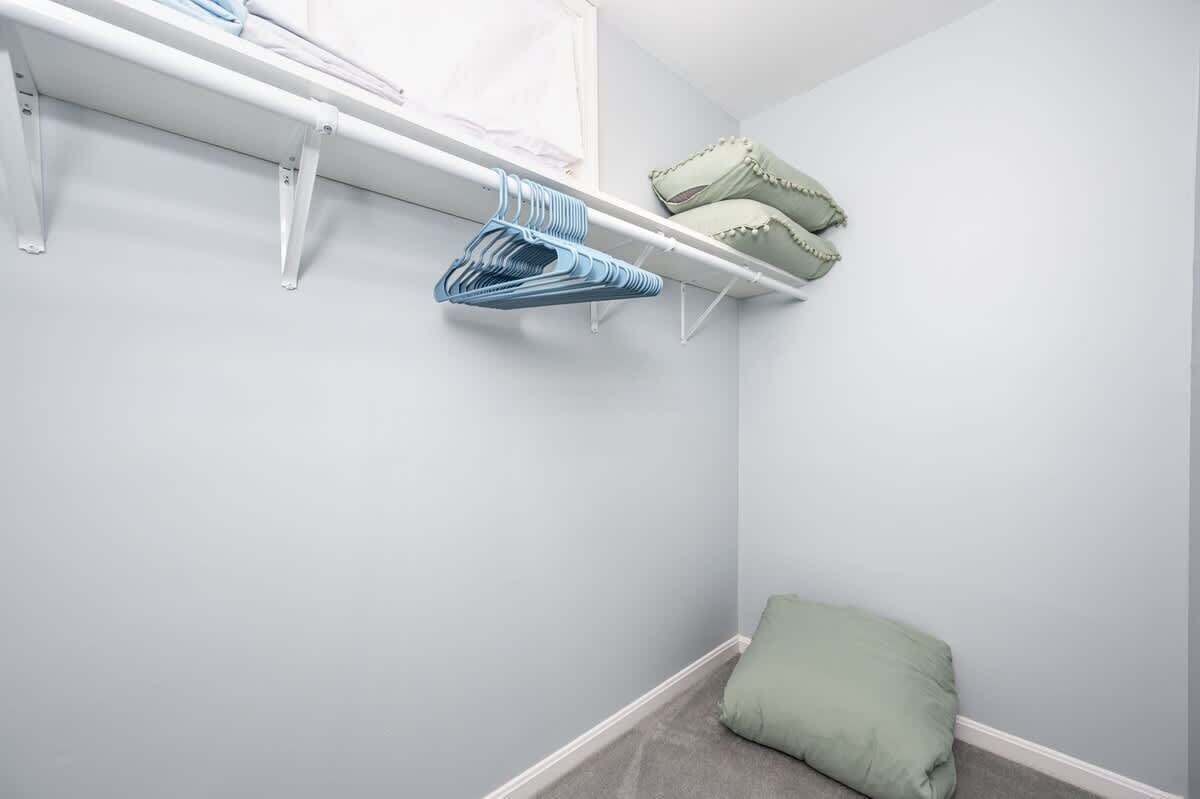 A corner of a room with a white shelf holding folded clothes and a green pillow on the floor.