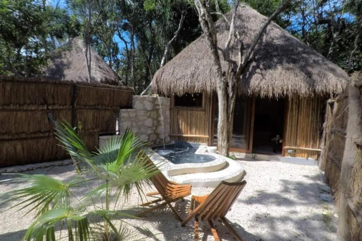 A thatched-roof hut with a stone wall, a small pool, and wooden furniture in a natural setting.