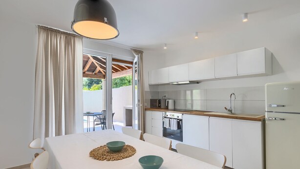 A modern kitchen with white cabinets, a built-in oven, and a refrigerator. There is a dining table with chairs and a large window with curtains.
