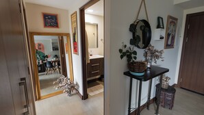 A hallway with a wooden door, a mirror, and a small table with a plant. There is a framed picture on the wall and a small stool with decorative items.