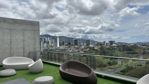 A rooftop terrace with modern seating, a concrete wall, and a view of a cityscape with buildings and mountains.