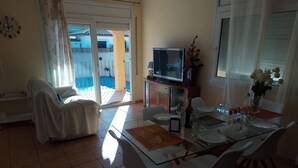 Ein Wohnzimmer mit einer Couch, einem Esstisch für zwei Personen, einem Fernseher und einem Balkon mit Blick auf einen Pool.
