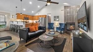 Un salon moderne avec un canapé en cuir noir, deux fauteuils, une table basse et une cheminée. On aperçoit la cuisine à l’arrière-plan, avec des meubles en bois et un four à micro-ondes.