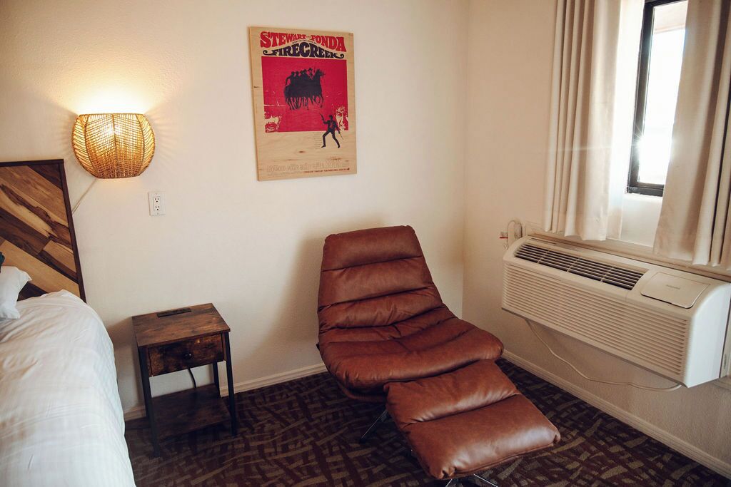Een hotelkamer met een bed, een bruine leren fauteuil, een klein houten nachtkastje, een wandlamp, een poster en een airconditioning.