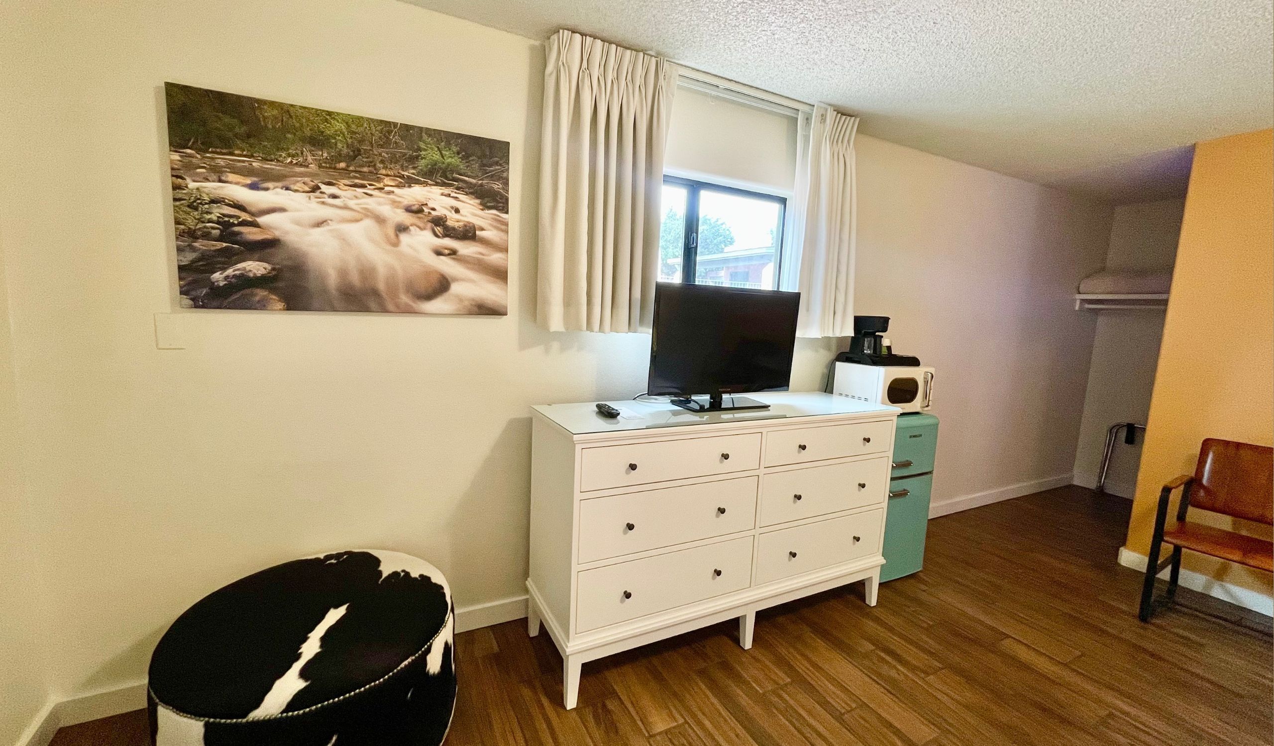 A room with a white dresser, a television on top, a window with curtains, and a painting on the wall.
